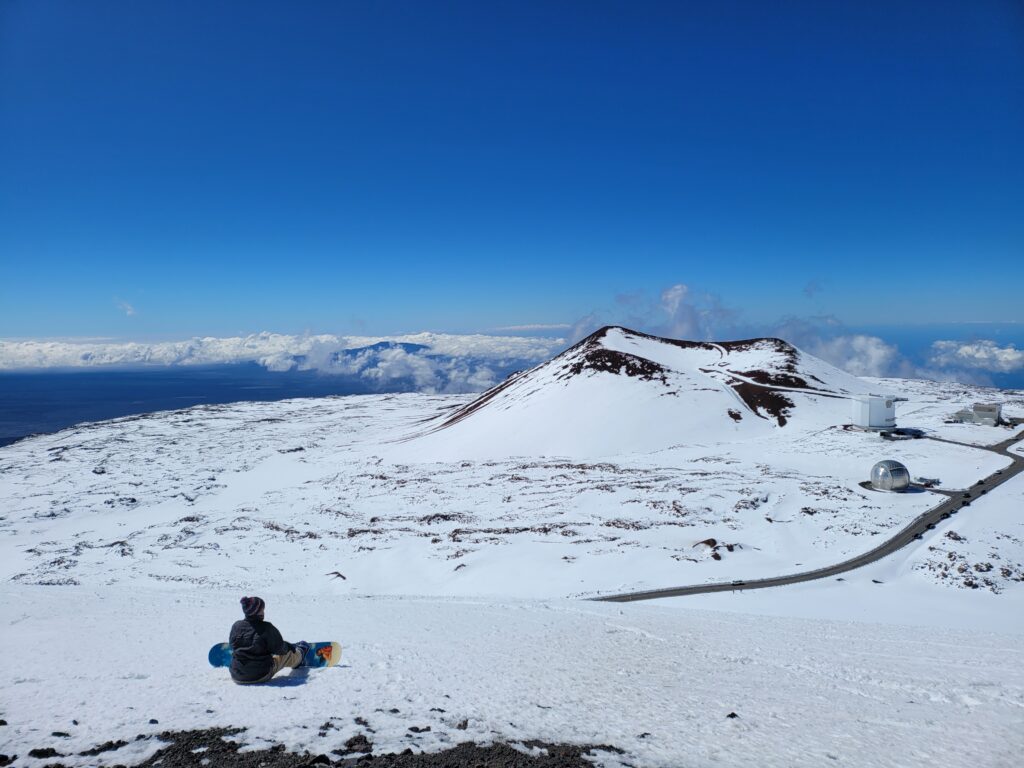A snowboarder takes a break and soaks in the view of a snowy Hawaii atop Mauna Kea. Maui appears in the distance with the clouds. Image: Weatherboy