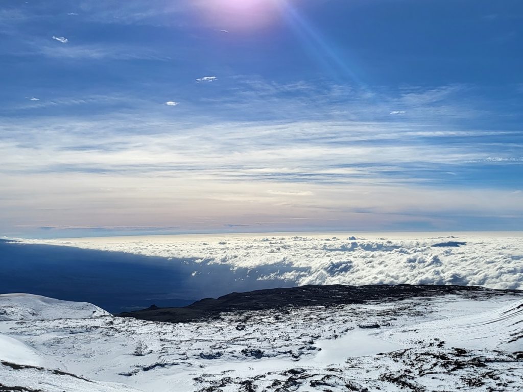 The view south towards Kona shows clouds locked-in below the peak of Mauna Loa as far as the eye can see. In the foreground, snow-covered slopes blend in with the nearby white clouds. Image: Weatherboy