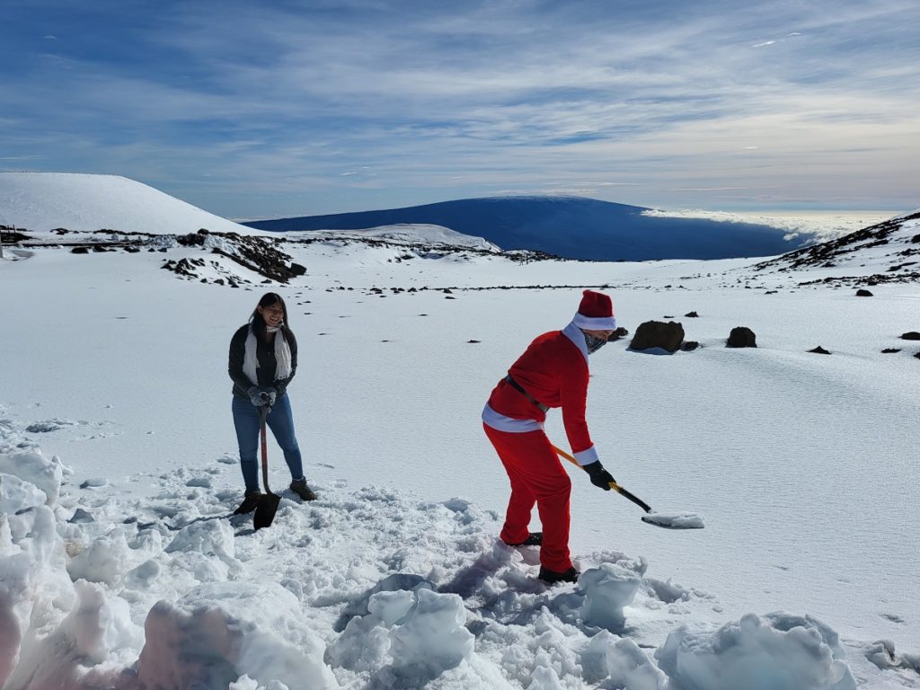 Big Island residents Reed from Kona, dressed as Santa, and Carrie from Hilo, shovel snow into a cooler near the summit of Mauna Kea. They are bringing snow to their friends near the beach in Kona to have a snowball fight with. Image: Weatherboy