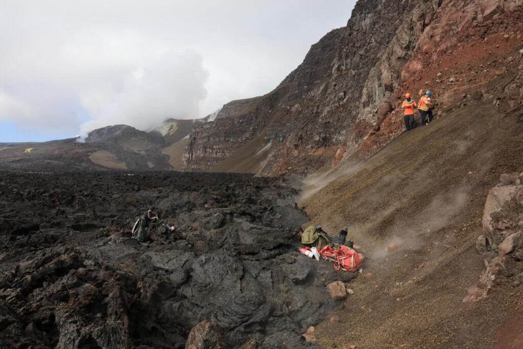 On July 2, three USGS Hawaiian Volcano Observatory scientists flew via helicopter to the northwest side of Halema‘uma‘u at the summit of Kīlauea to sample tephra deposits in the crater wall. These deposits were exposed by the 2018 summit collapse, but lava flows from the ongoing episodic Kīlauea summit eruption are likely to bury them again in the near future. Refilling the of the crater by eruptions in Halema‘uma‘u since 2020, including the ongoing episode lava fountaining eruption made it fairly easy to access the deposits, which just a few years ago were halfway up the cliff. The vent complex from the ongoing eruption can be seen in the distance on the left. Image: USGS / M. Zoeller